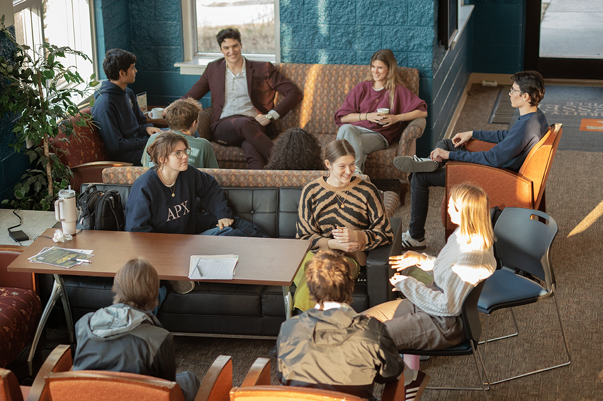 A group of 11 students sit couches and chairs talking.