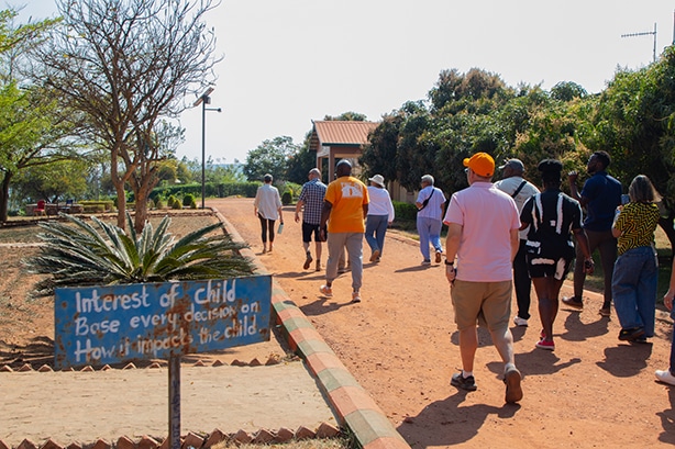 A group of people walk along a red-dirt path toward a small building, while a hand-painted sign in the foreground reads, “Interest of child: Base every decision on how it impacts the child.”