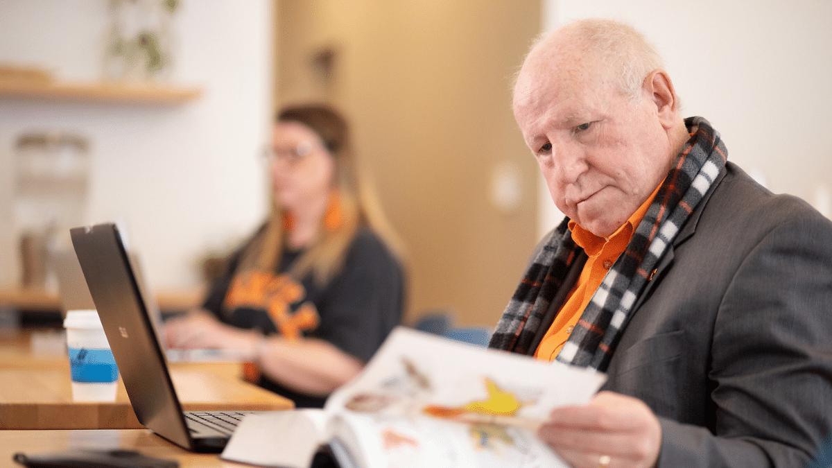 Man looking at a textbook while he's on his laptop.