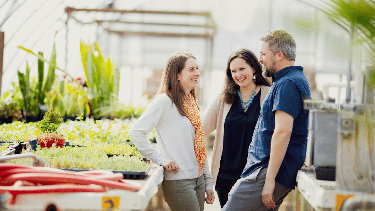 Two women and a man chatting inside a greenhouse.